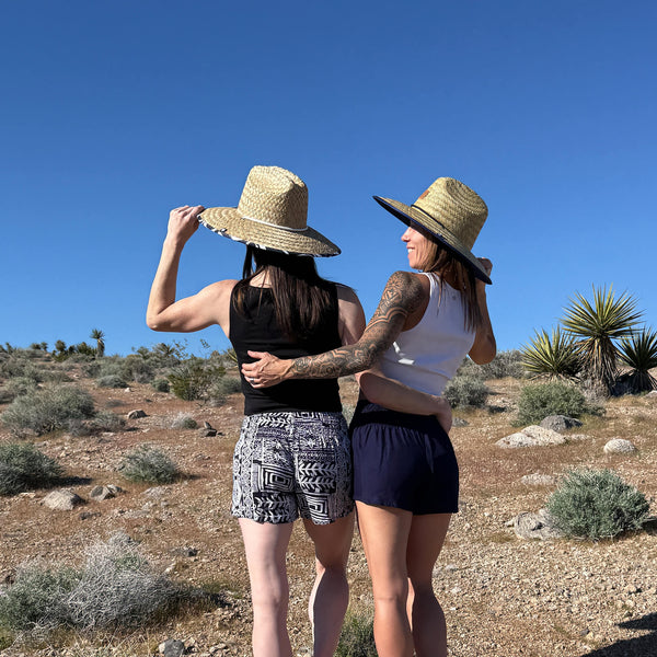 Back view of two women in ocean wave shorts with straw hats — Ember Glyph boho shorts